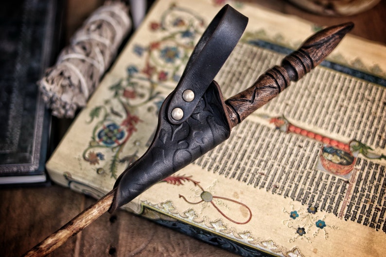 Close up of a black embossed leather wand holster holding a carved wooden wand, resting on an illuminated medieval manuscript decorated with floral borders. The holster loop shows two rounded rivets and the leather texture appears aged and shaped by hand. Perfect accessory for wizard cosplay, witch costume, fantasy mages and larp wand holder gear. The scene includes bundled sage and old books, reinforcing the magical, alchemist inspired atmosphere.