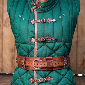 May include: A sleeveless green quilted gambeson vest featuring a high collar with brown leather trim, three front leather buckle closures, and a wide brown leather belt with a metal buckle. The garment displays the text Outcast Props.