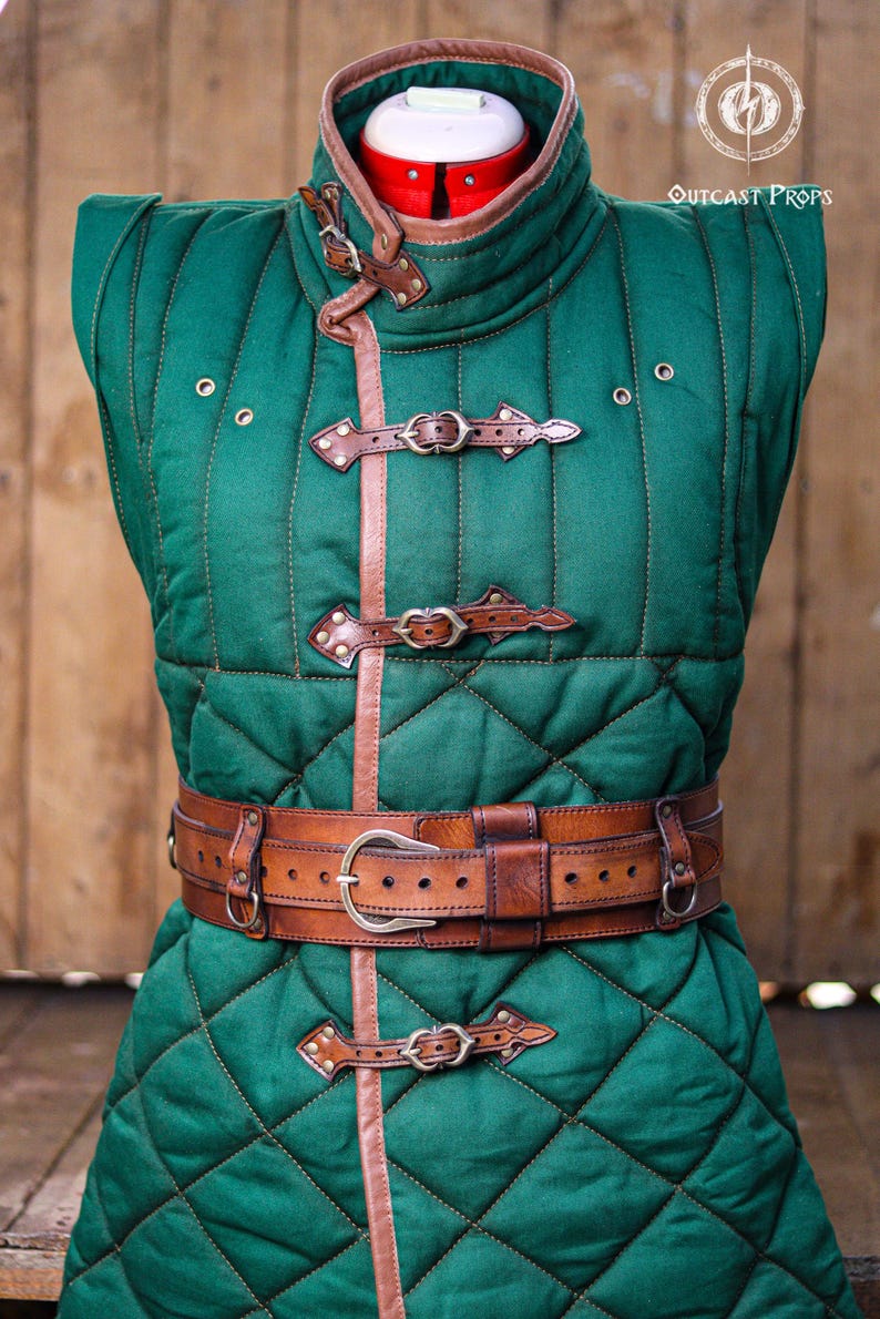 May include: A sleeveless green quilted gambeson vest featuring a high collar with brown leather trim, three front leather buckle closures, and a wide brown leather belt with a metal buckle. The garment displays the text Outcast Props.