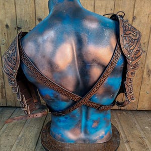 Rear view of Viking leather shoulder armor Sedrik, showcasing adjustable leather straps crossed on the back of a sculpted mannequin torso. Detailed engraved Celtic patterns on leather pauldrons visible from both sides. Rustic wooden background enhances the handcrafted leather's aged aesthetic. Ideal for Norse cosplay, Celtic warrior costume, Viking LARP gear, fantasy armor set, medieval reenactment outfit, premium leather shoulder armor.