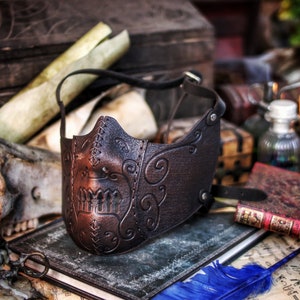 May include: A brown leather mask with intricate floral designs and a stitched edge. The mask has a skull-like shape with a raised, textured design on the front.