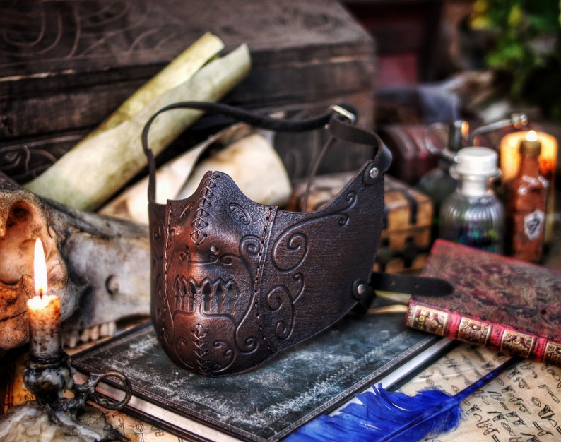 May include: A brown leather mask with intricate floral designs and a stitched edge. The mask has a skull-like shape with a raised, textured design on the front.