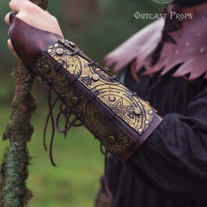 Close-up of golden demon hunter leather bracer worn on a male arm, showing tied leather cords and engraved symbols of protection. Ideal for LARP spellcasters, cultist costumes or witch cosplay. Made from durable leather, dyed and painted by hand for fantasy realism. A comfortable, adjustable accessory that combines dark mysticism with functional design.