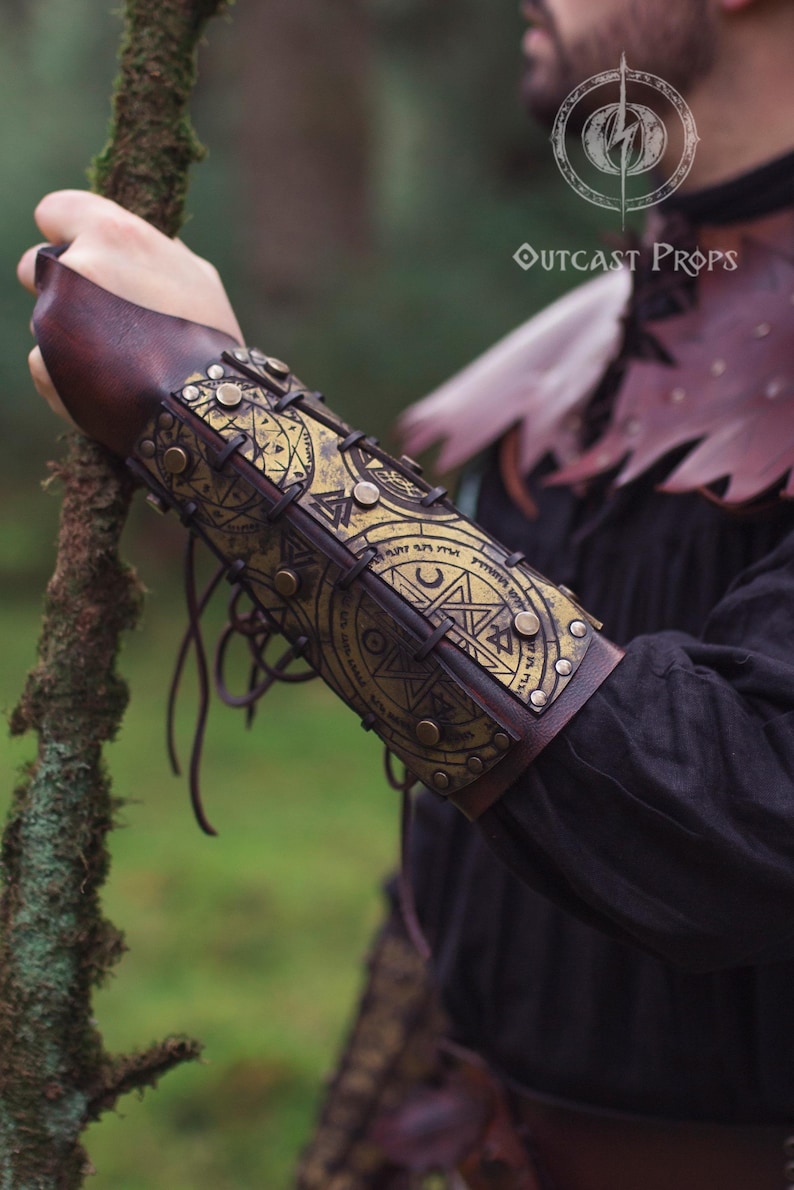 Close-up of golden demon hunter leather bracer worn on a male arm, showing tied leather cords and engraved symbols of protection. Ideal for LARP spellcasters, cultist costumes or witch cosplay. Made from durable leather, dyed and painted by hand for fantasy realism. A comfortable, adjustable accessory that combines dark mysticism with functional design.