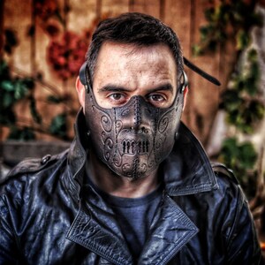 May include: A person wearing a black leather jacket and a brown leather mask with intricate designs. The mask covers the person's mouth and nose and has a stitched seam down the center.