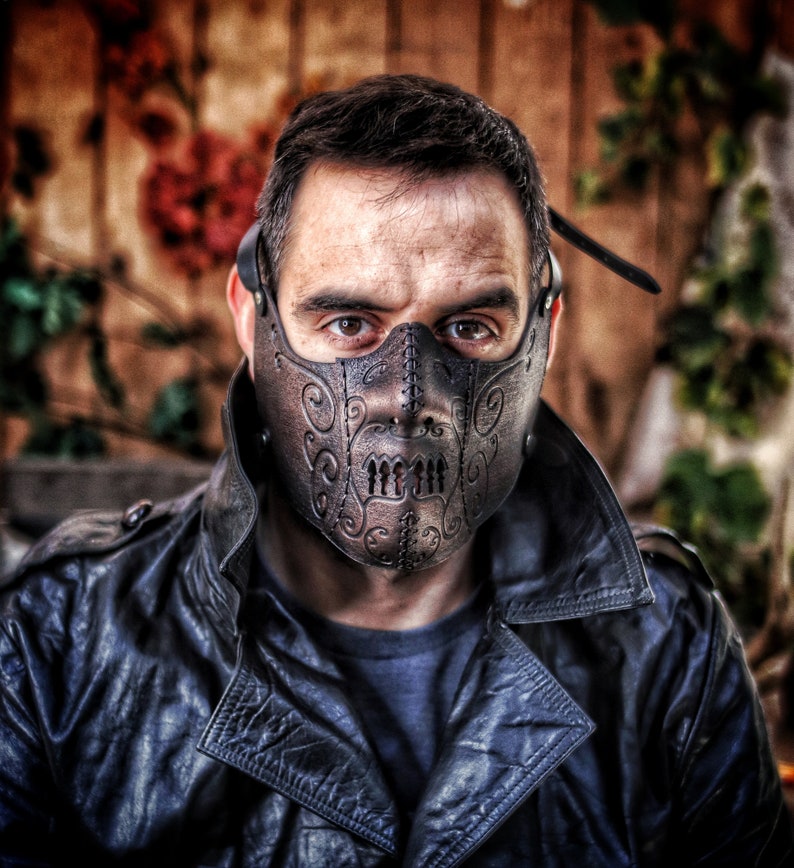May include: A person wearing a black leather jacket and a brown leather mask with intricate designs. The mask covers the person's mouth and nose and has a stitched seam down the center.
