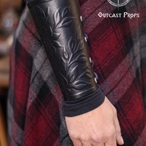 Close up of a black floral leather bracer worn over a long sleeve shirt and paired with a plaid renaissance skirt. The renaissance faire bracer shows embossed leaves and petals that form a refined floral arm guard. Ideal as an elf cosplay accessory, fae costume accessory or larp bracer, the piece highlights artisan embossing and smooth shaping that flatters the arm, perfect for fantasy or medieval inspired outfits. Fae costume, ren faire, druid larp, leaf design, floral design.