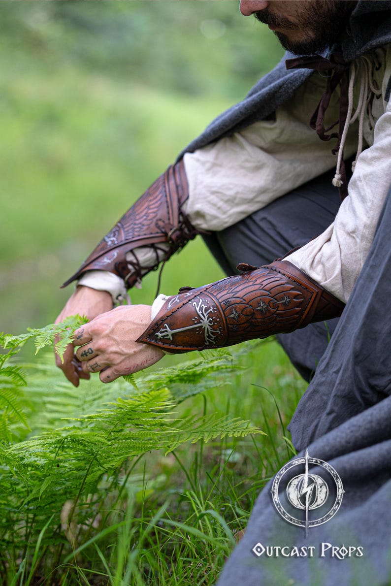 Elven ranger kneeling in nature, collecting herbs while wearing brown embossed vambraces with White Tree and stars. Handcrafted accessory ideal for LOTR cosplay, LARP or portraying Aragorn, Boromir, or Faramir. Perfect for Tolkien-inspired reenactments, medieval fantasy fairs or ranger costumes. Designed for flexibility and realism in fantasy roleplay. Made with aged vegetable-tanned leather. White tree leather bracers fantasy ranger armor LARP bracers medieval cosplay accessory elven warrior