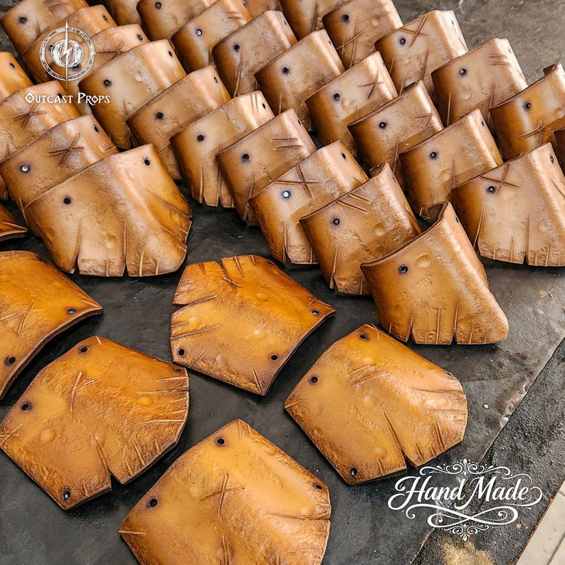 May include: A collection of handcrafted, weathered leather armor pieces. The pieces are various shades of brown with visible stitching and rivet details. The Outcast Props logo is visible in the upper left corner, and the words "Hand Made" are in the lower right.