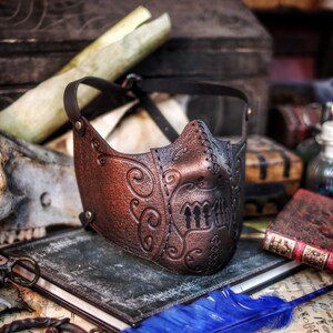 May include: A brown leather mask with intricate floral designs and a stitched seam. The mask has a dark brown leather strap and is decorated with a pattern of three vertical lines.