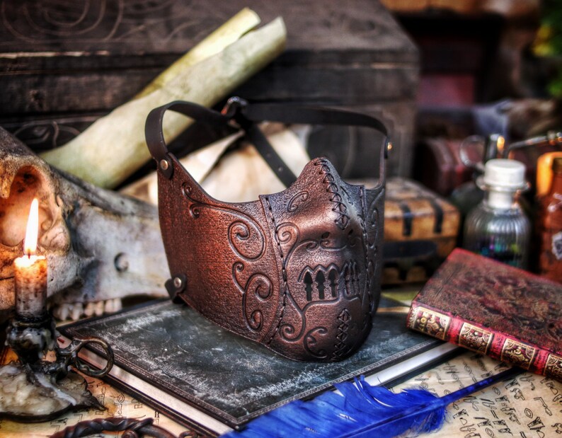 May include: A brown leather mask with intricate floral designs and a stitched seam. The mask has a dark brown leather strap and is decorated with a pattern of three vertical lines.