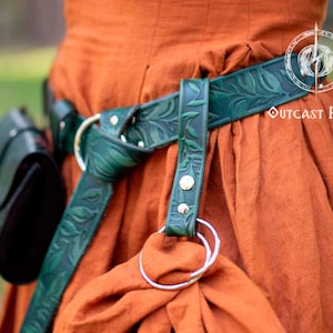 Può includere: Una cintura in pelle verde con un motivo floreale, indossata su un abito color ruggine. La cintura ha una fibbia argentata e un anello argentato attaccato.