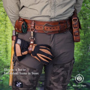 May include: A brown leather utility belt featuring a metal buckle, a small green pouch, a metal clip holding a black and tan glove, and a side holster. A wooden tool handle is visible on the left side. Text reads: Shop as a Set or Individual Items in Store.