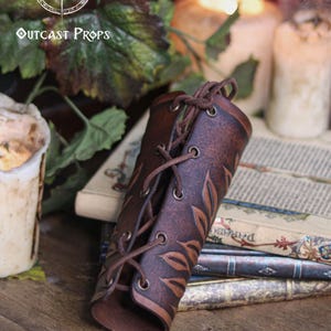 A single aged brown floral leather bracer lies partially rolled on top of antique books beside melted candles and ivy leaves. This renaissance faire bracer features embossed botanical patterns and laced fastening, creating a delicate floral arm guard. Ideal as an elf cosplay accessory, fae costume accessory or larp bracer, the handcrafted texture suits romantic fantasy and woodland inspired characters. Fae costume, ren faire, druid larp, leaf design, floral design.