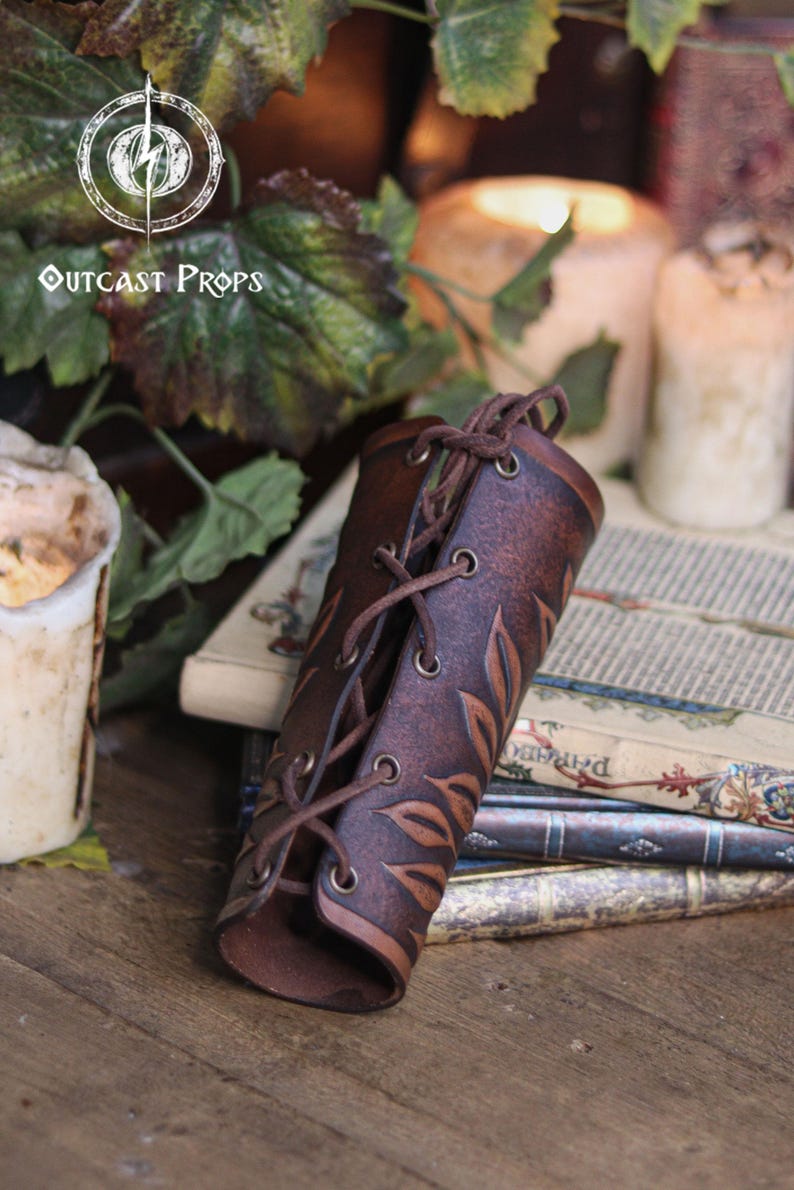 A single aged brown floral leather bracer lies partially rolled on top of antique books beside melted candles and ivy leaves. This renaissance faire bracer features embossed botanical patterns and laced fastening, creating a delicate floral arm guard. Ideal as an elf cosplay accessory, fae costume accessory or larp bracer, the handcrafted texture suits romantic fantasy and woodland inspired characters. Fae costume, ren faire, druid larp, leaf design, floral design.