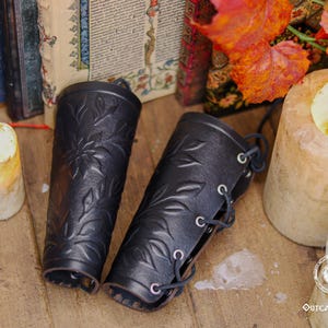 A pair of black floral leather bracers rests beside candles and old books in a rustic fantasy setting. Each renaissance faire bracer features deep embossed leaves and flower motifs, offering a romantic handcrafted look. Ideal as an elf cosplay accessory, floral arm guard or larp bracer, this fae costume accessory suits renaissance faire outfits, woodland characters or herbalist themed costumes. Fae costume, ren faire, druid larp, leaf design, floral design.