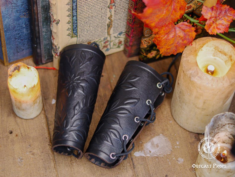 A pair of black floral leather bracers rests beside candles and old books in a rustic fantasy setting. Each renaissance faire bracer features deep embossed leaves and flower motifs, offering a romantic handcrafted look. Ideal as an elf cosplay accessory, floral arm guard or larp bracer, this fae costume accessory suits renaissance faire outfits, woodland characters or herbalist themed costumes. Fae costume, ren faire, druid larp, leaf design, floral design.