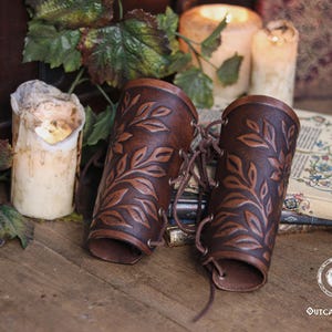 Two aged brown floral leather bracers with laced fastening lay on antique books surrounded by ivy and candlelight. The renaissance faire bracer design highlights natural shading and embossed leaves, creating a handcrafted floral arm guard ideal as an elf cosplay accessory or fae costume accessory. The rustic scene enhances their larp bracer appeal for herbalists, forest characters and medieval fantasy costumes. Fae costume, ren faire, druid larp, leaf design, floral design.