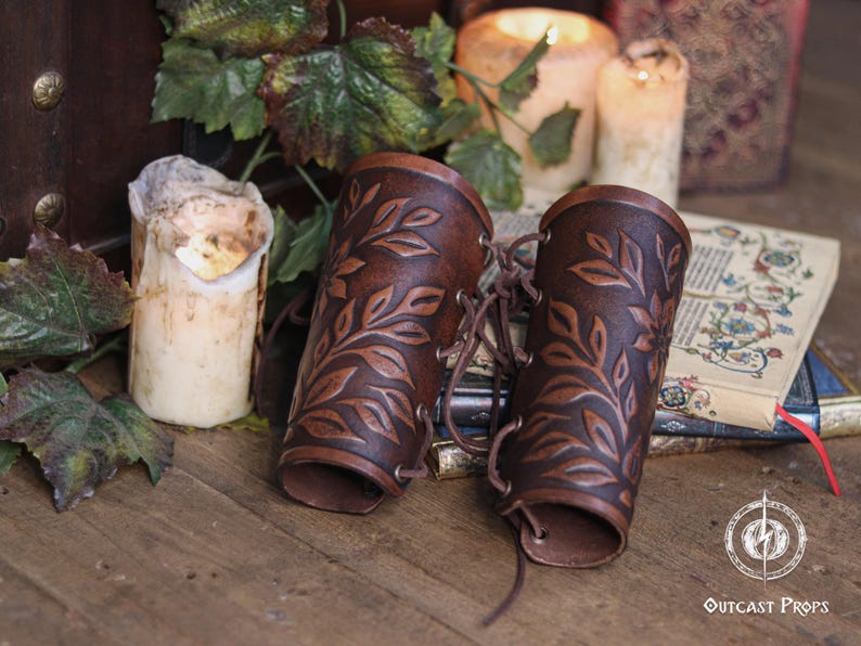 Two aged brown floral leather bracers with laced fastening lay on antique books surrounded by ivy and candlelight. The renaissance faire bracer design highlights natural shading and embossed leaves, creating a handcrafted floral arm guard ideal as an elf cosplay accessory or fae costume accessory. The rustic scene enhances their larp bracer appeal for herbalists, forest characters and medieval fantasy costumes. Fae costume, ren faire, druid larp, leaf design, floral design.