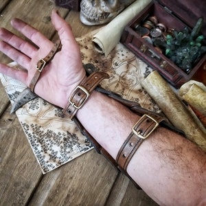 Close-up of a male arm wearing an open Dragon Scales Leather Bracer, showing the inner leather straps and antique brass buckles used to fasten it. The bracer lies on a rustic wooden table with medieval props like a map, coins, scrolls and a skull. Ideal for LARP warrior costume, fantasy fair outfits or orc archer cosplay. This handcrafted bracer is made of premium vegetable-tanned leather using traditional Spanish techniques.