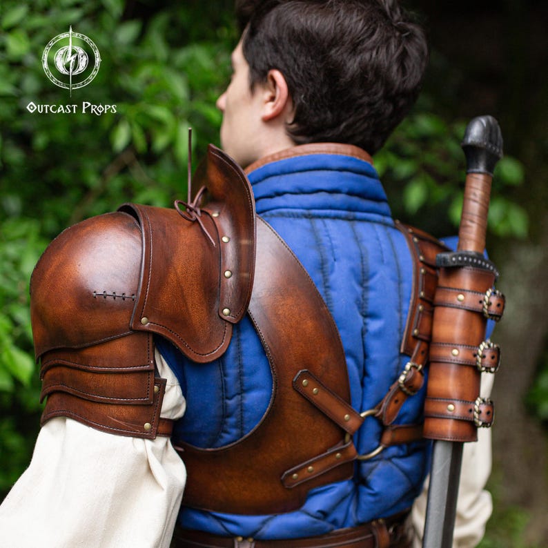 Pu&ograve; includere: Armatura per spalle in pelle marrone con dettagli rivettati e un fodero per spada abbinato. L'armatura &egrave; indossata sopra un capo trapuntato blu. L'immagine presenta il logo "Outcast Props" nell'angolo in alto a sinistra.