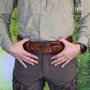 May include: A wide brown leather belt featuring a metal buckle, multiple adjustment holes, and integrated leather loops. The belt is worn over a light olive button-down shirt and dark brown trousers, with a logo visible in the upper right corner.