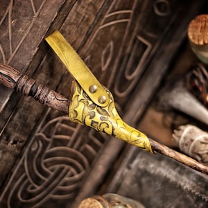 A yellow embossed leather wand holster holds a carved wooden wand in a rustic fantasy setting. The holster hangs against a carved wooden chest with Celtic style patterns, surrounded by sage, bottles and parchment. Its aged dye finish and swirling embossing stand out vividly. The scene evokes alchemist gear, wizard cosplay accessories, witch costume details and renaissance faire magic wand holster style.