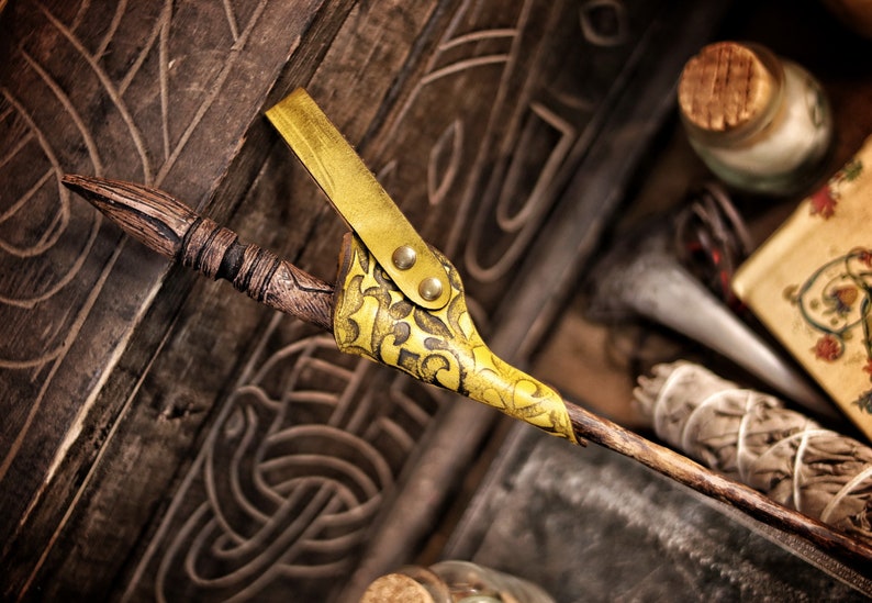 A yellow embossed leather wand holster holds a carved wooden wand in a rustic fantasy setting. The holster hangs against a carved wooden chest with Celtic style patterns, surrounded by sage, bottles and parchment. Its aged dye finish and swirling embossing stand out vividly. The scene evokes alchemist gear, wizard cosplay accessories, witch costume details and renaissance faire magic wand holster style.