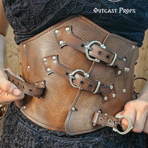Front detail of the aged brown leather underbust corset in nickel finish, handcrafted with three decorative buckle straps and hidden clasp insert. Designed for witches and fantasy characters, this steampunk waist cincher suits LARP, gothic, and renaissance outfits. Vegetable-tanned leather aged with unique techniques ensures a durable, authentic look. Adjustable with leather side and back laces for comfort and precision.