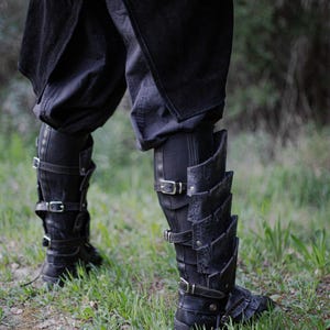 May include: Black leather leg armor with multiple buckles and layered plates. The armor is worn over black pants and boots. The image is taken outdoors with green grass in the background. The Outcast Props logo is visible.