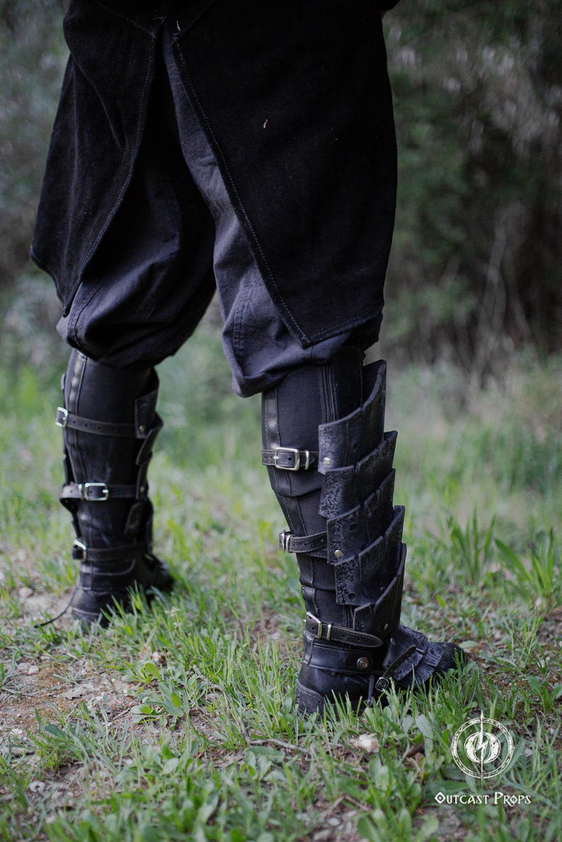 May include: Black leather leg armor with multiple buckles and layered plates. The armor is worn over black pants and boots. The image is taken outdoors with green grass in the background. The Outcast Props logo is visible.