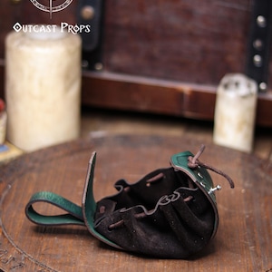 A green leather coin pouch is shown open on a wooden surface, revealing its soft dark suede interior with gathered folds and a drawstring closure. The structured leather flap features botanical embossing, while the back loop allows it to hang from a belt. In the blurred background there is a wooden chest and candles, creating a fantasy workshop atmosphere. This elven herbalist renfaire pouch is ideal for coins, dice or small items at a renaissance faire or LARP.