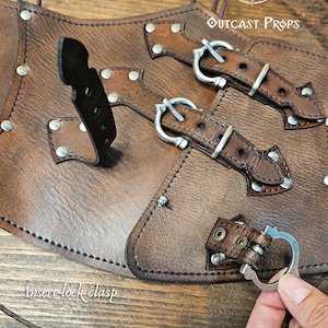Detailed image of the aged brown leather corset with nickel buckles showing the insert-lock clasp system on a wooden Outcast Props display. The handcrafted underbust corset features triple front straps for closure and alignment. Perfect for steampunk, ren faire, and witch costume looks, this piece highlights Outcast Props’ artisanal work and eco-friendly vegetable-tanned leather finish.