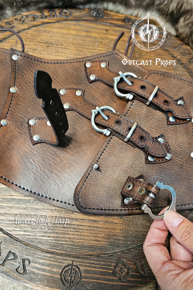 Detailed image of the aged brown leather corset with nickel buckles showing the insert-lock clasp system on a wooden Outcast Props display. The handcrafted underbust corset features triple front straps for closure and alignment. Perfect for steampunk, ren faire, and witch costume looks, this piece highlights Outcast Props’ artisanal work and eco-friendly vegetable-tanned leather finish.