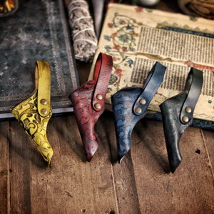 Four colorful leather wand holsters in yellow, red, blue and green rest side by side on a wooden table near an antique manuscript. Each holster features embossed swirling motifs, riveted belt loops and a distinct hand dyed finish. This lineup highlights customization options for wizard cosplay, witch costume, larp wand holder accessories and renaissance faire outfits. The aged fantasy setting emphasizes their handcrafted artisan quality.