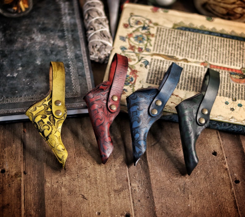 Four colorful leather wand holsters in yellow, red, blue and green rest side by side on a wooden table near an antique manuscript. Each holster features embossed swirling motifs, riveted belt loops and a distinct hand dyed finish. This lineup highlights customization options for wizard cosplay, witch costume, larp wand holder accessories and renaissance faire outfits. The aged fantasy setting emphasizes their handcrafted artisan quality.