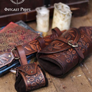 A brown floral embossed leather coin pouch sits beside a large matching hip bag and belt, displayed on wooden floorboards with candles and ornate books in the background. Text Belt and hipbag not included is shown. The pouch’s suede body and botanical carving make it ideal for elves, witches, herbalists and renfaire nobles. Perfect as a leather coin bag or fantasy dice pouch for renaissance faire outfits, LARP or immersive cosplay.