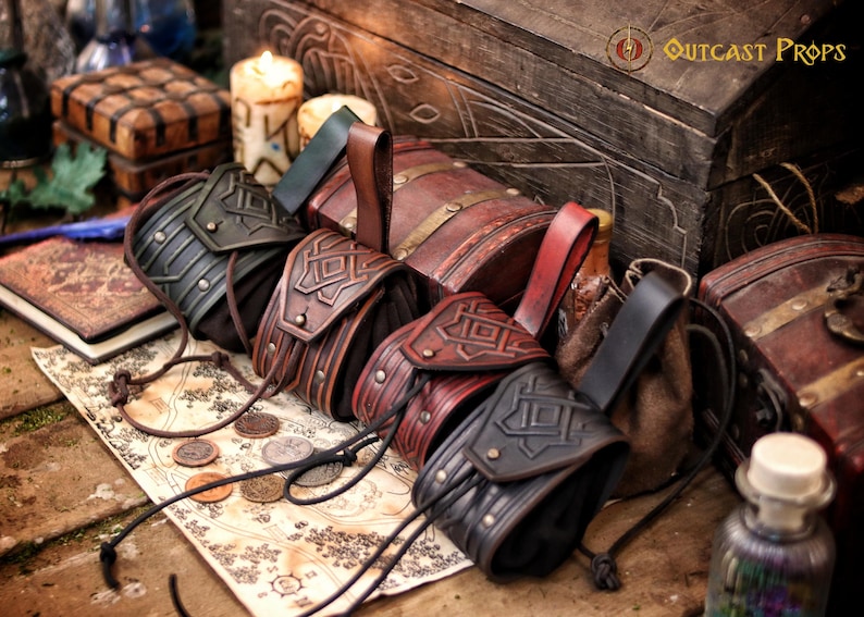 Group of handcrafted leather coin pouches in green, brown, red and black leather, arranged on an illustrated fantasy map with metal coins. Each pouch is made from vegetable-tanned leather with embossed geometric patterns, suede sides and adjustable drawstrings. The belt loops allow secure wearing during LARP, medieval reenactment, Viking or Celtic costumes. The scene includes wooden chests, candles and books, creating a timeless fantasy atmosphere.
