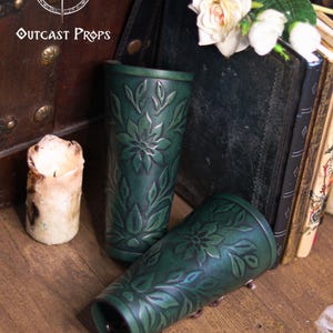 Aged green floral leather bracers shown in a rustic medieval setting with candles and old books. Each renaissance faire bracer displays engraved botanical patterns and smooth curved shaping for the arm. Suitable as an elf cosplay accessory, fae costume accessory, herbalist gear or larp bracer. The floral arm guard design blends artisan embossing with vibrant dye work for romantic fantasy costumes. Fae costume, ren faire, druid larp, leaf design, floral design.