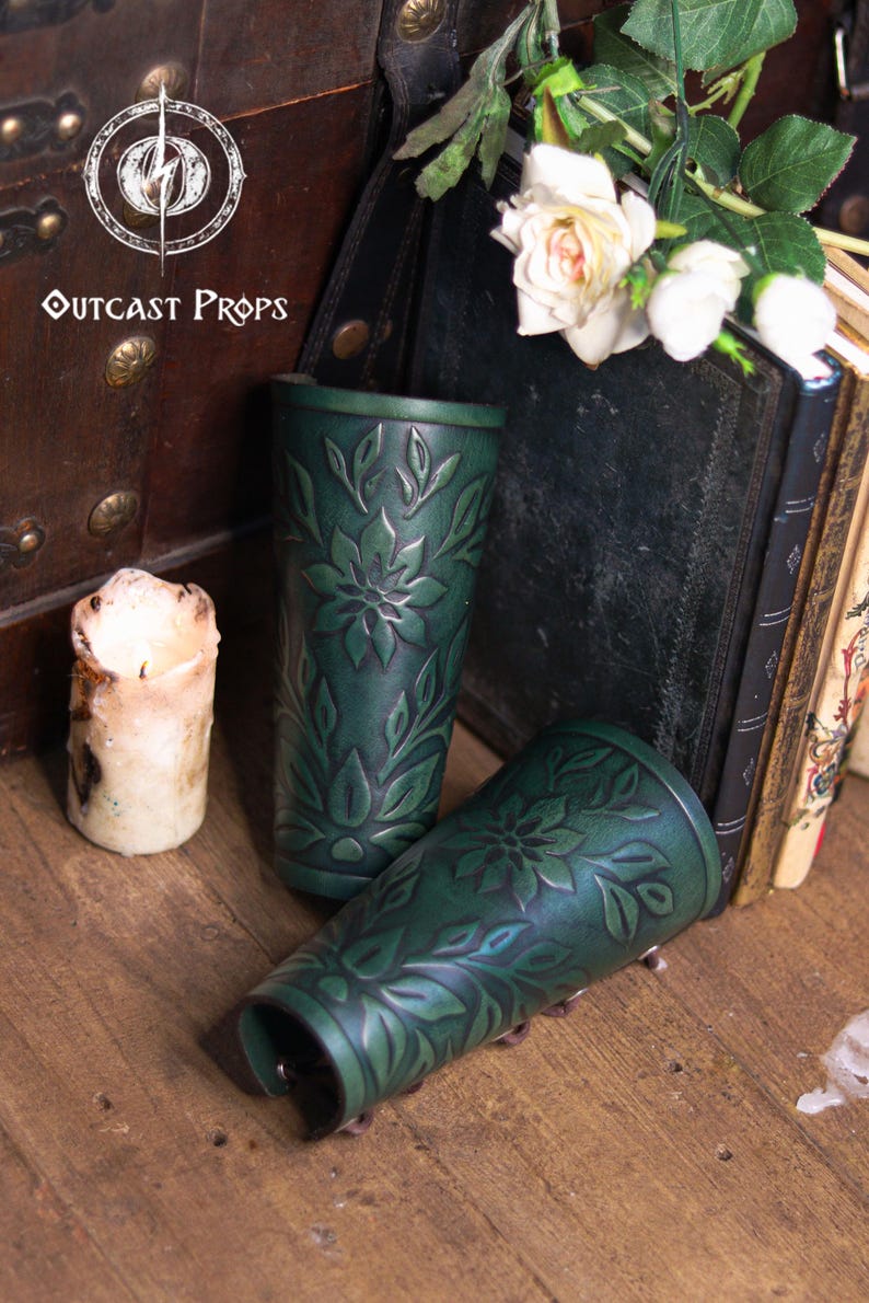 Aged green floral leather bracers shown in a rustic medieval setting with candles and old books. Each renaissance faire bracer displays engraved botanical patterns and smooth curved shaping for the arm. Suitable as an elf cosplay accessory, fae costume accessory, herbalist gear or larp bracer. The floral arm guard design blends artisan embossing with vibrant dye work for romantic fantasy costumes. Fae costume, ren faire, druid larp, leaf design, floral design.