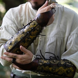 Close-up of a warrior holding a blade while wearing golden magic bracers engraved with alchemist and summoning sigils. The leather has a warm aged finish, laced with black cords and metal studs. Ideal for LARP, cosplay, or fantasy characters like demon hunters or arcane warriors. These ornate leather bracers add a mystical touch to any medieval or fantasy outfit, combining protection, storytelling, and artisanal detail. Demon Hunter Leather Bracers Armor for LARP Cosplay Witch Warlock Alchemist