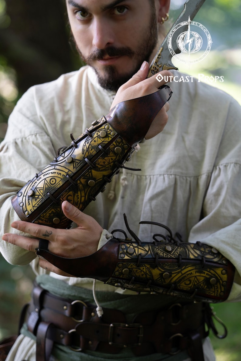 Close-up of a warrior holding a blade while wearing golden magic bracers engraved with alchemist and summoning sigils. The leather has a warm aged finish, laced with black cords and metal studs. Ideal for LARP, cosplay, or fantasy characters like demon hunters or arcane warriors. These ornate leather bracers add a mystical touch to any medieval or fantasy outfit, combining protection, storytelling, and artisanal detail. Demon Hunter Leather Bracers Armor for LARP Cosplay Witch Warlock Alchemist