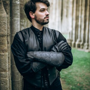 Man dressed in full black medieval outfit poses confidently with arms crossed, showcasing detailed black leather bracers embossed with Yggdrasil, the Norse tree of life. These vambraces are ideal for LARP armor, viking cosplay or as part of a Geralt witcher costume. The setting of ancient stone pillars and green grass adds an immersive backdrop for fantasy characters. Great for larping, female warrior armor, or medieval leather accessories.