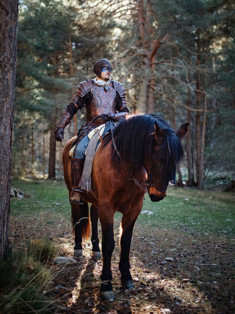 May include: A person wearing leather armor and a helmet with blue face paint rides a brown horse through a forest. The horse has a black mane and tail.