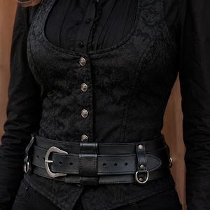 Black bushcraft leather belt worn over a black gothic vest and shirt, showing a wide utility belt design with inner adjustable strap, outer leather frame, nickel hardware and side attachment rings. Handmade vegetable tanned leather belt for daily wear, steampunk outfit, LARP costume, tactical style, military inspired look, post apocalyptic character, ren faire gear and medieval fantasy clothing. The belt creates a strong waist silhouette while remaining practical for modular pouches