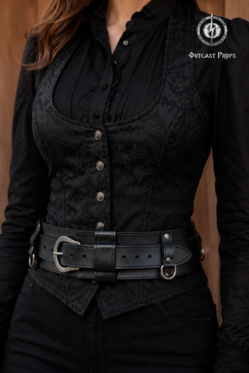 Black bushcraft leather belt worn over a black gothic vest and shirt, showing a wide utility belt design with inner adjustable strap, outer leather frame, nickel hardware and side attachment rings. Handmade vegetable tanned leather belt for daily wear, steampunk outfit, LARP costume, tactical style, military inspired look, post apocalyptic character, ren faire gear and medieval fantasy clothing. The belt creates a strong waist silhouette while remaining practical for modular pouches