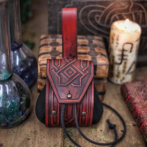 Close-up of a red leather coin pouch with a triangular embossed flap and visible belt loop, photographed against a wooden chest and candlelit background. The pouch is crafted from premium vegetable-tanned leather, hand dyed and aged to enhance the carved geometric lines. The suede body allows flexible storage for coins while maintaining a compact shape. This leather coin pouch is suitable for LARP characters, Viking or Celtic costumes, dwarf or adventurer roles, Renaissance fairs and fantasy