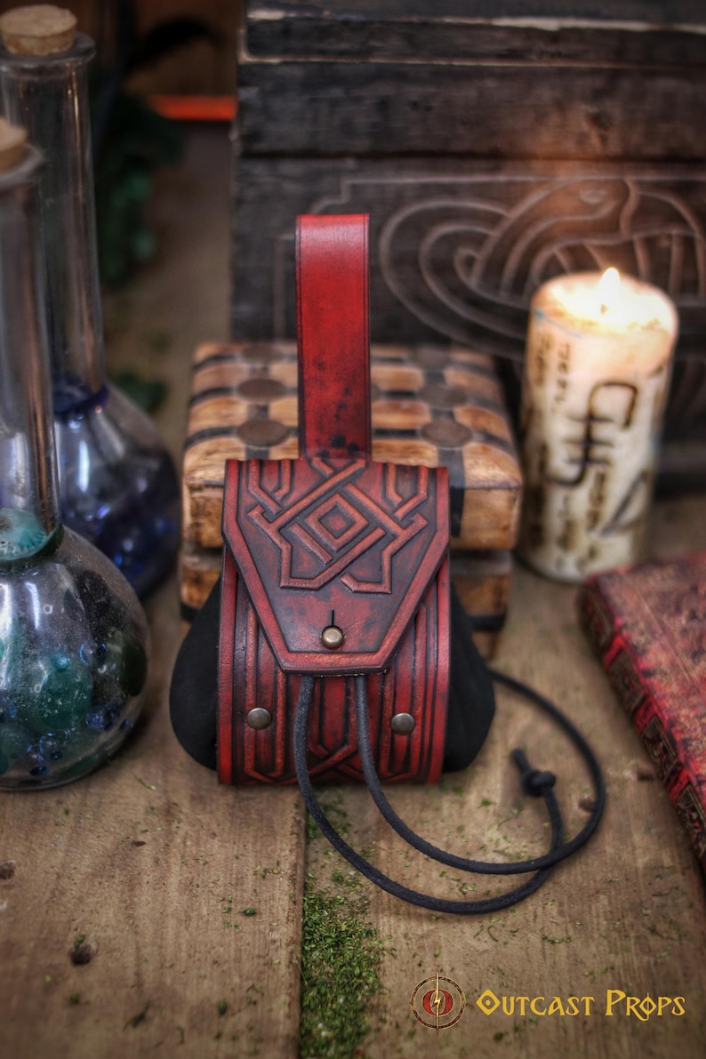 Close-up of a red leather coin pouch with a triangular embossed flap and visible belt loop, photographed against a wooden chest and candlelit background. The pouch is crafted from premium vegetable-tanned leather, hand dyed and aged to enhance the carved geometric lines. The suede body allows flexible storage for coins while maintaining a compact shape. This leather coin pouch is suitable for LARP characters, Viking or Celtic costumes, dwarf or adventurer roles, Renaissance fairs and fantasy