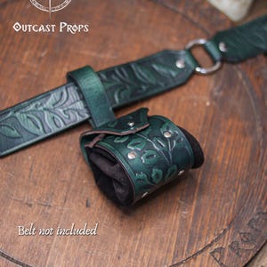 A detailed view of a green floral leather coin pouch attached to a matching embossed belt, shown against a wooden background. The coin bag combines botanical embossing, metal rivets and suede sides, creating an elegant accessory for elven characters, witches, druids or herbalists. Perfect for renfaire and renaissance faire outfits, it holds coins, dice or keys securely as a small fantasy pouch for LARP and immersive costume wear.