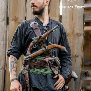 May include: A person wearing a black shirt and grey trousers, with a leather belt and two antique pistols. The pistols are holstered on a brown leather harness. The image also includes a metal mug and a knife. The text "Cutcast Props" is visible.